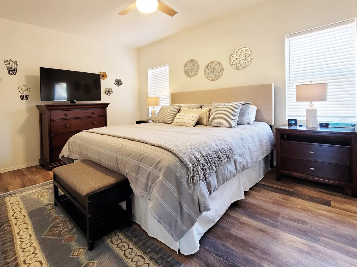 Here, a cozy bedroom is arranged with a bed topped with decorative pillows, a dresser with a TV, nightstands with lamps, and stylish wall art. The room’s soft lighting and peaceful mood make it an ideal sanctuary for relaxation.