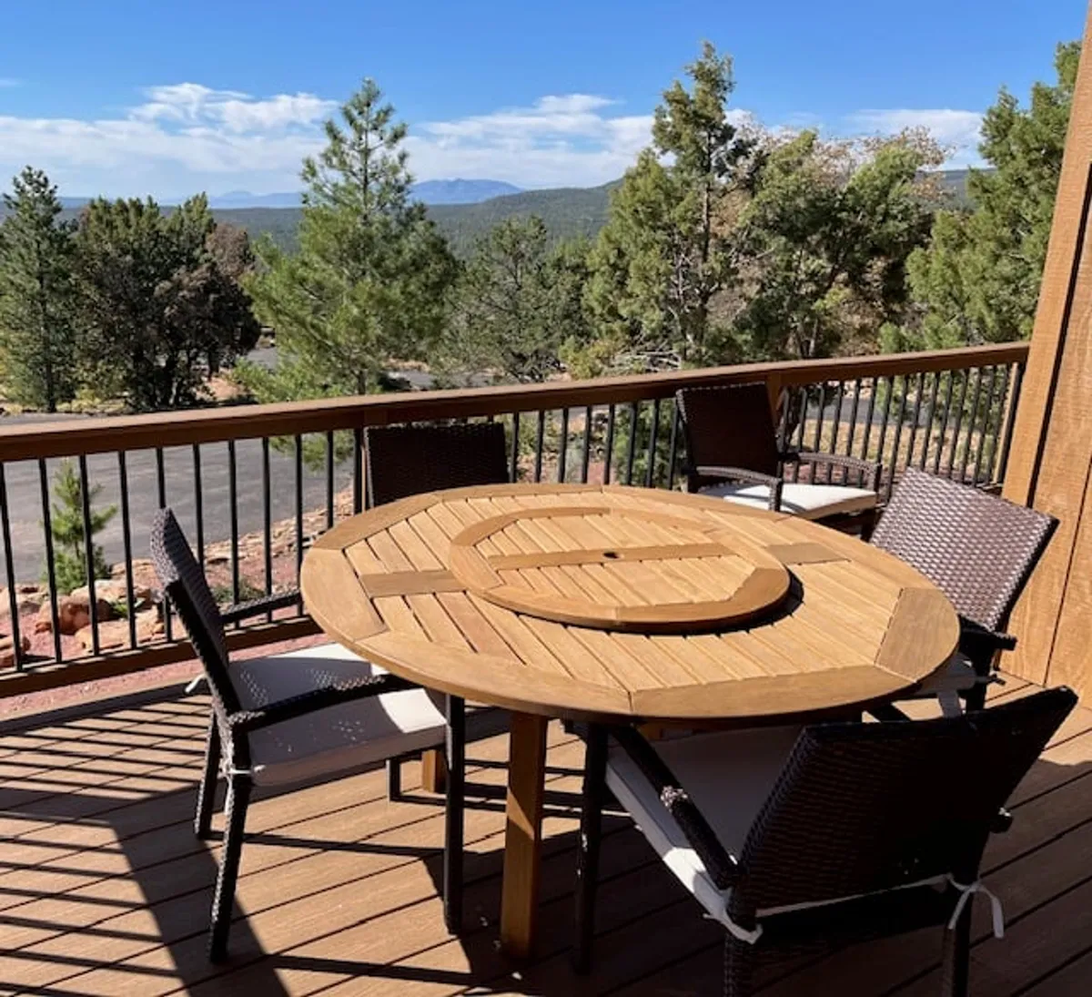 A wooden round table with surrounding chairs sits on a deck, nestled among lush greenery with distant mountain views. The scene is serene, evoking the idea of sharing morning coffee or evening drinks while enjoying the tranquil mountain surroundings. The setting is intimate, perfect for couples seeking quiet moments together.