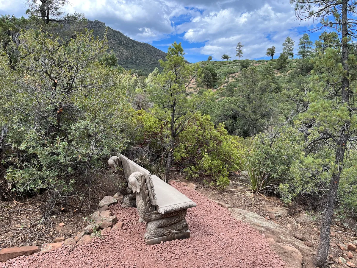A peaceful outdoor scene featuring an ornate stone bench set amidst lush greenery, with a breathtaking mountainous backdrop. The environment feels serene and inviting, perfect for quiet moments in nature.