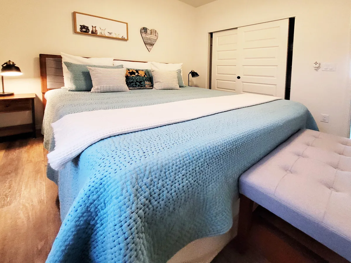 This image showcases a spacious bedroom featuring a large bed with a textured blanket, decorative pillows, a nightstand with a lamp, framed art on the wall, and a bench at the foot of the bed. Soft lighting and calming décor create an atmosphere of relaxation and comfort.
