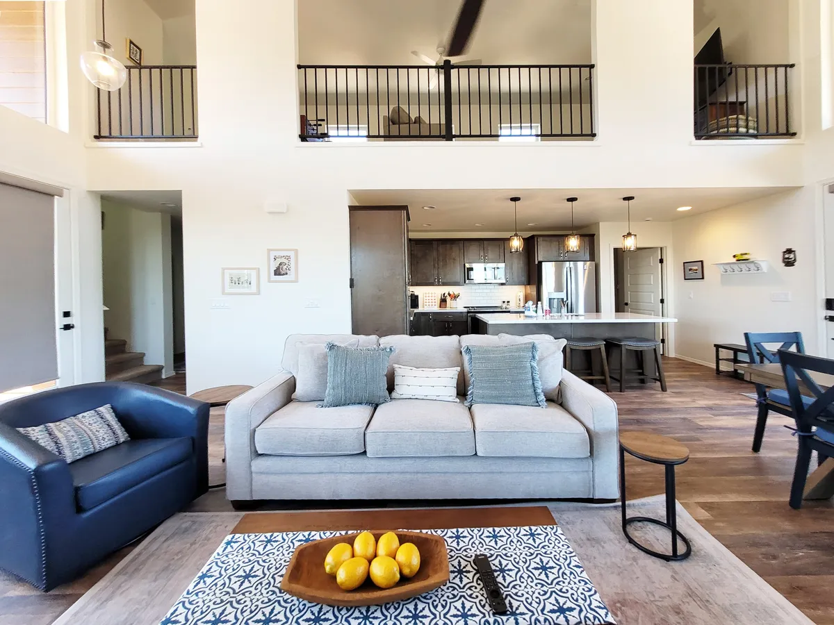 This image presents an open-concept area where the kitchen seamlessly connects to a cozy living space. A sofa with decorative pillows sits beside a coffee table with fruit, and the kitchen area is visible in the background, suggesting a flow between cooking, dining, and relaxation.