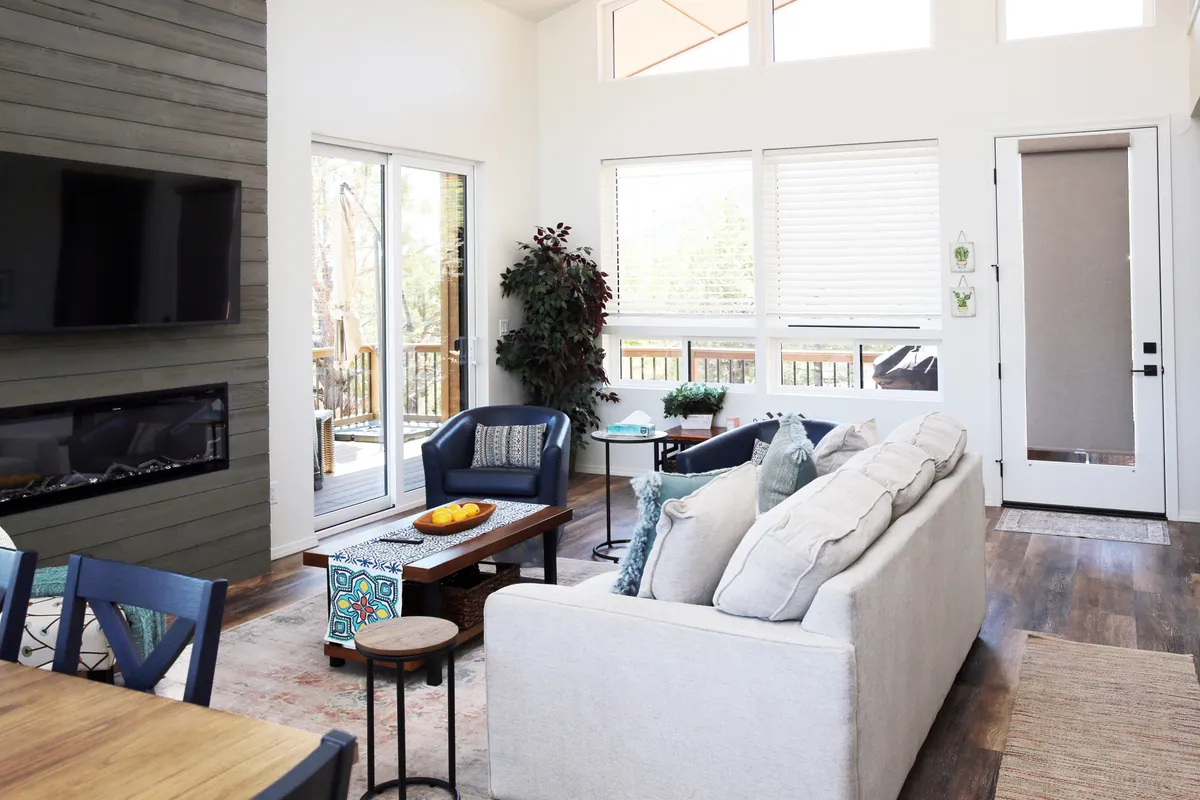A bright, stylish living room is shown with a plush sofa, coffee table, plant accents, a fireplace, and expansive windows that draw the outdoors in. The setting feels tranquil yet sociable, inviting both relaxation and conversation.