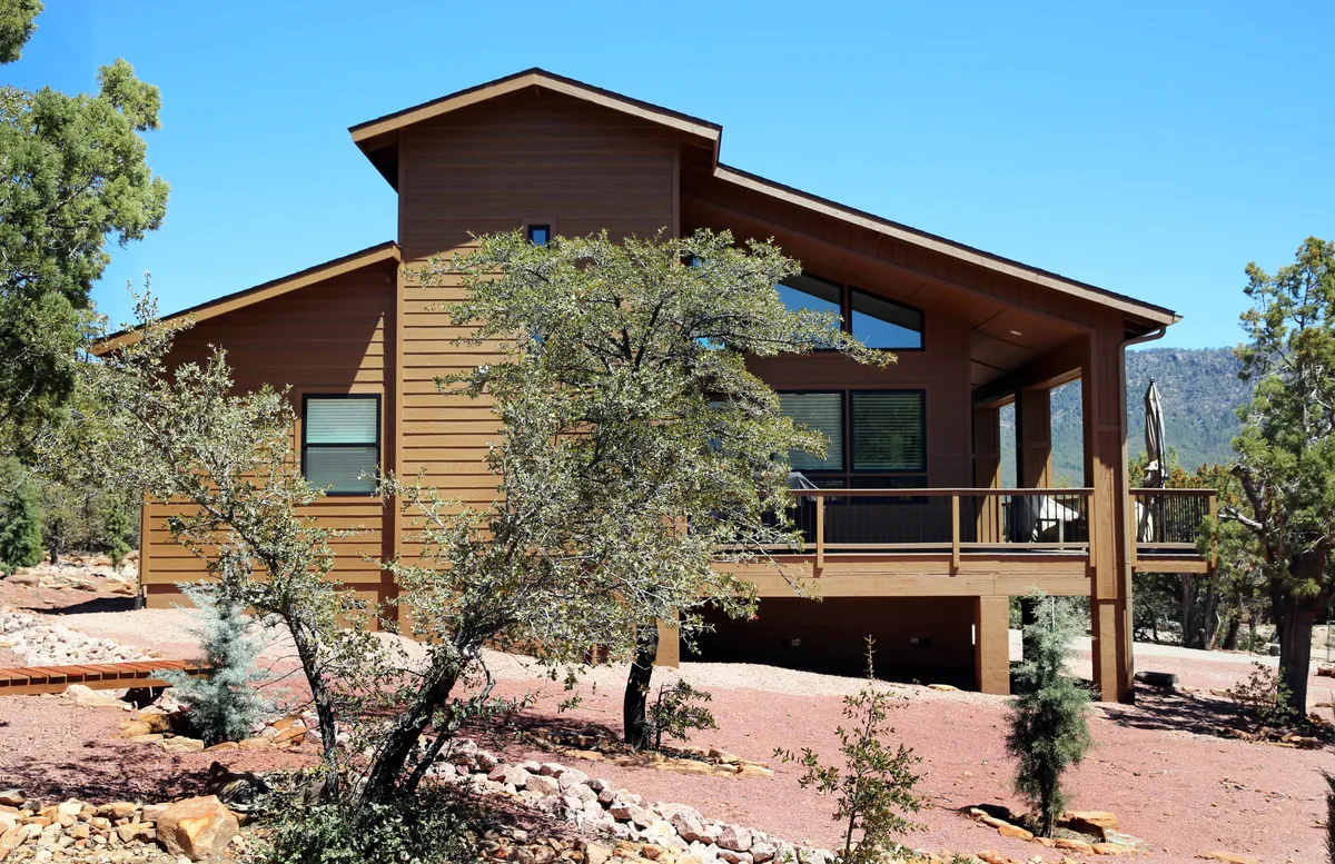 The modern wooden exterior of the house features large windows and an expansive deck area. Guests are seen relaxing on the deck, enjoying the transition between indoor comfort and the beauty of the surrounding nature, with hints of sunset colors enhancing the mood.